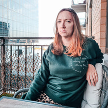 Load image into Gallery viewer, Woman with long blonde hair and green eyes sitting on a new York city apartment balcony looking towards the camera wearing a graphic print Green sweatshirt with white writing on top right chest area reads "i form calming circles with my dog" surrounded by two half circles of paws