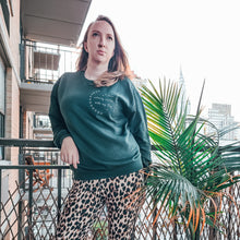 Load image into Gallery viewer, Woman with long blonde hair and green eyes standing on a new York city apartment balcony next to a plant looking up at the sky wearing leopard leggings and a Green dog mom graphic sweatshirt with white writing on top right chest area reads "i form calming circles with my dog" surrounded by two half circles of paws